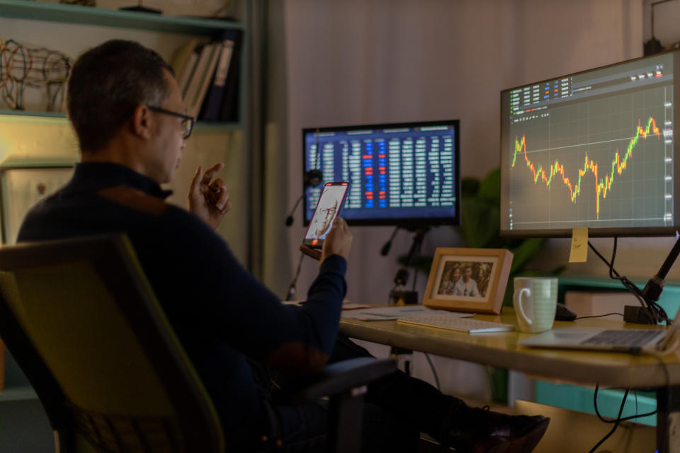 Man looking at his phone and two computer screens with data charts on them