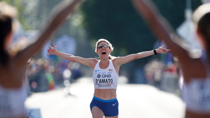 A woman with blonde hair and a white singlet celebrates as finishes the marathon