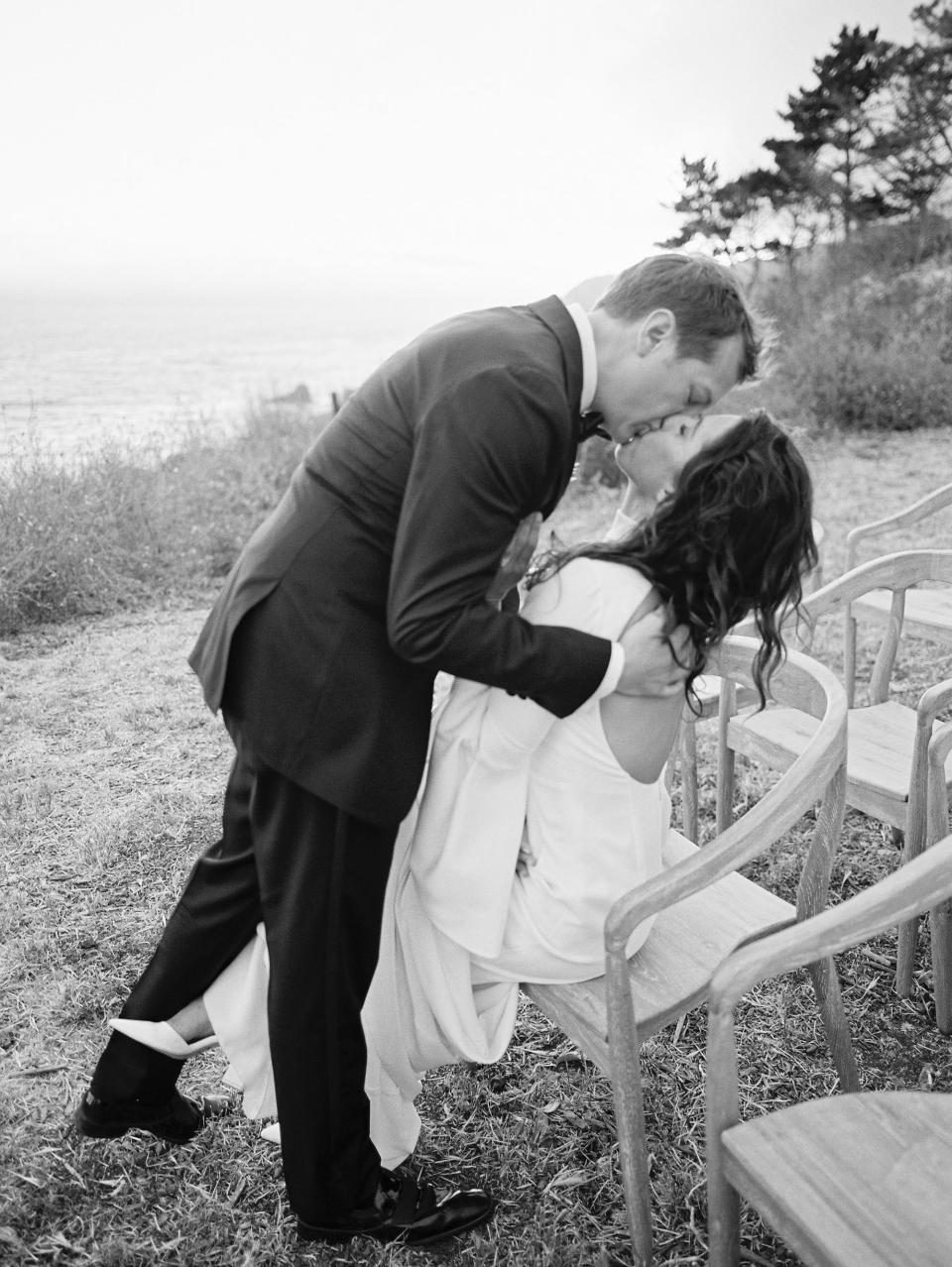 A groom leans down to kiss a bride who is sitting in a chair in black and white.