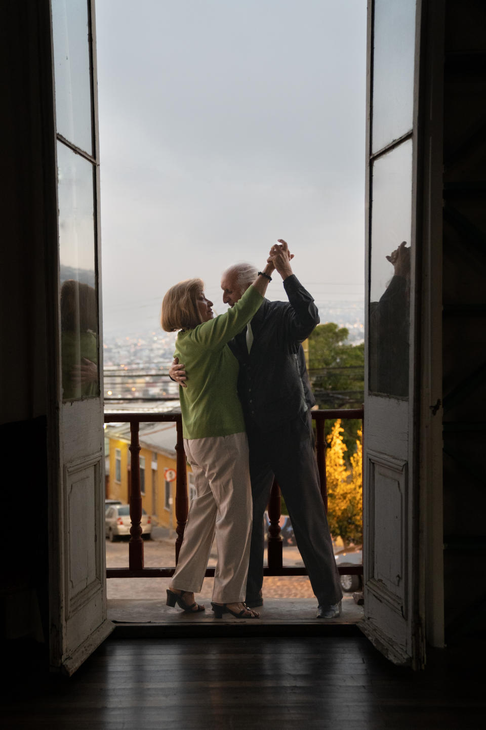 An elderly couple holds hands while dancing in front of a large, open doorway with a scenic view of a town and distant landscape behind them