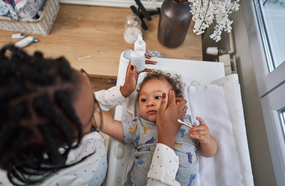 Caring single mother applying skin lotion on her baby boy while he lying on a change bed in his nursery