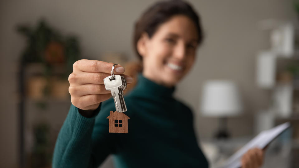 A woman is smiling while holding out a set of keys, one of which has a small house-shaped keychain. She holds a document in her other hand