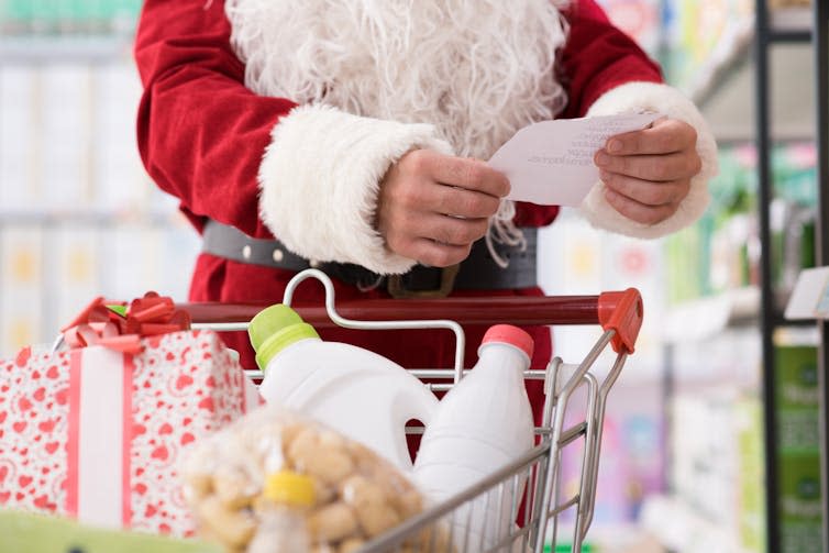 Torso and hands of a man in a Santa Claus costume, checking a grocery list while pushing a trolley in a supermarket