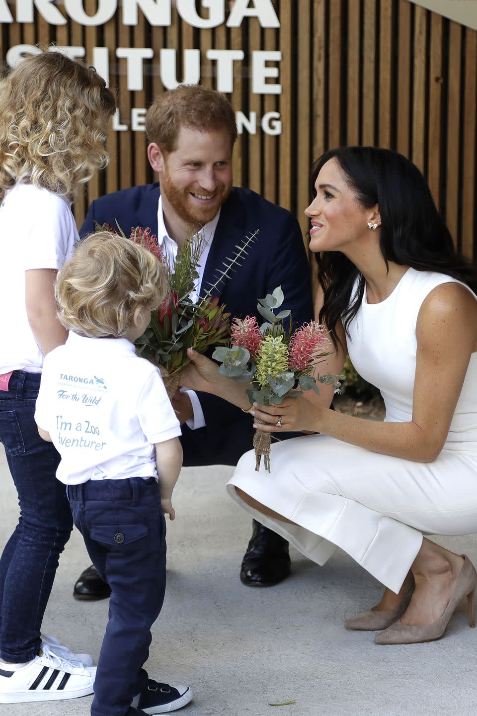 <p>Meghan receives some flowers. She is wearing a <a rel="nofollow noopener" href="https://www.harpersbazaar.com/fashion/designers/a23810788/meghan-markles-karen-gee-dress/" target="_blank" data-ylk="slk:white dress by Karen Gee;elm:context_link;itc:0;sec:content-canvas" class="link ">white dress by Karen Gee</a>.</p>