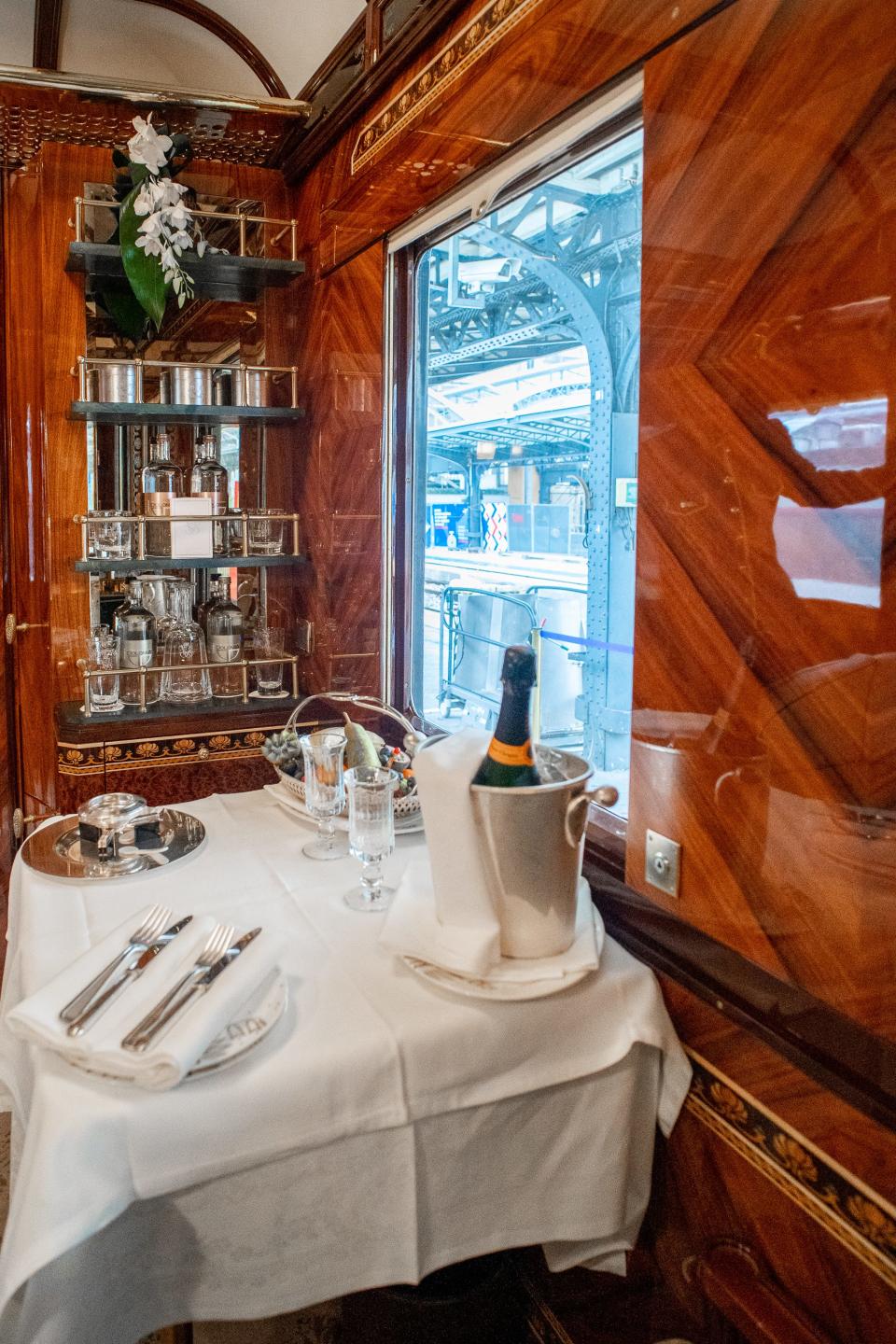 Dining table with white cloth and champagne in front of a bar on the wall in a train cabin with wooden walls