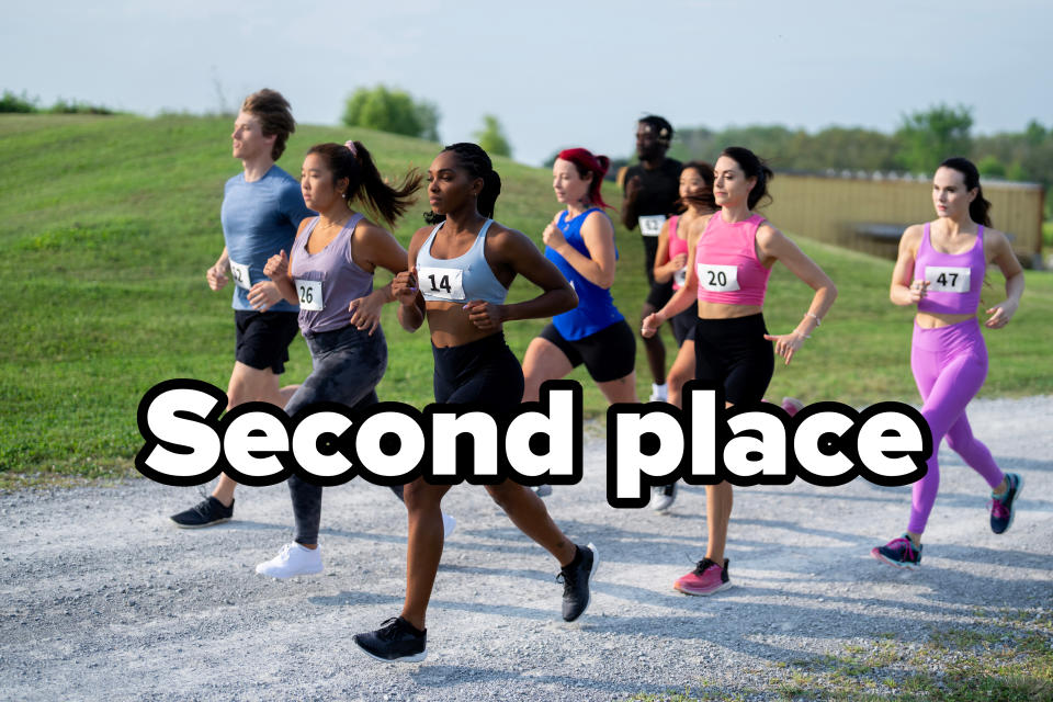 A group of runners, including numbered participants, race on a gravel path in an outdoor setting. The runners wear various athletic outfits and appear focused