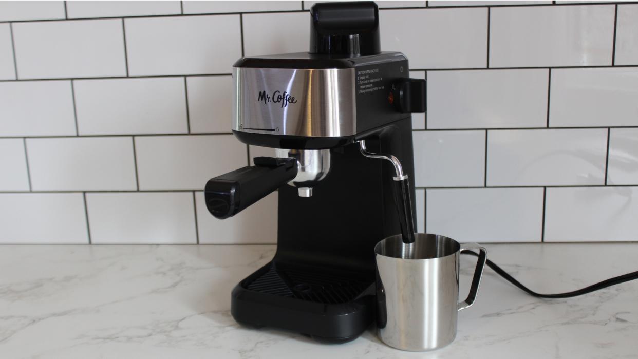  Mr. Coffee Steam Espresso Maker with stainless steel metal milk frothing pitcher on marble kitchen countertop with white tiled kitchen wall in background. 