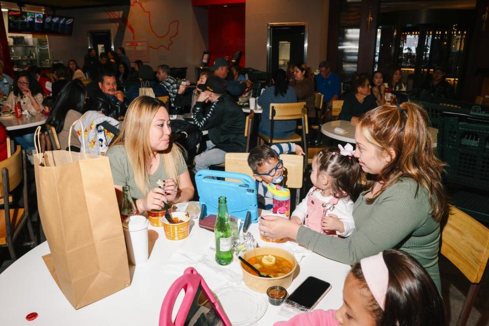 Two women and three children seated around a round white table in the middle of a food court.