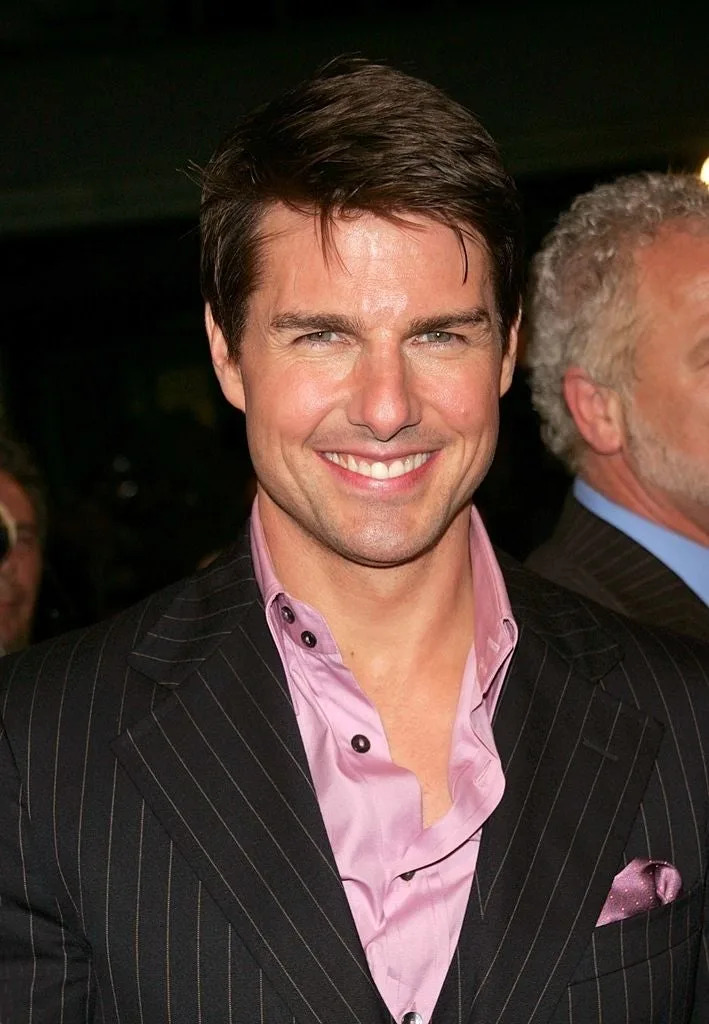 Tom Cruise in a pinstripe suit and pink shirt, smiling at an event. A man with white hair is partially visible behind him