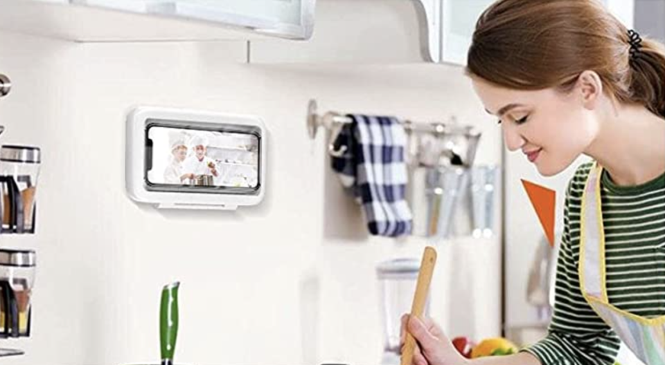 A dark-haired woman stirs a pot in the kitchen while her shower phone holder is visible on the wall above.