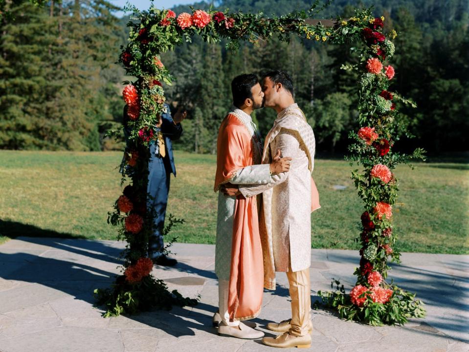 Two grooms kiss under a floral arch in front of a blue sky.