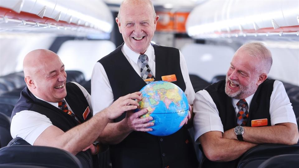 Three older male cabin crew members are pictured aboard an easyJet aircraft. One of them is holding a globe while his colleague points to a country on the map.