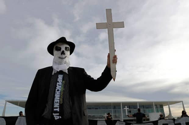 A demonstrator dressed as a ghost and wearing a tie calling for the impeachment of President Jair Bolsonaro holds a cross representing more than 300,000 COVID-19-related deaths outside the Planalto presidential palace in Brasilia, Brazil, Friday, March 26, 2021.