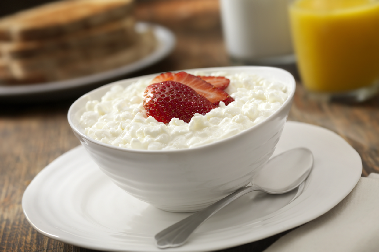 Cottage cheese in a white bowl and matching white plate with a spoon in the foreground and peanut butter toast on a white plate, blurred in the background