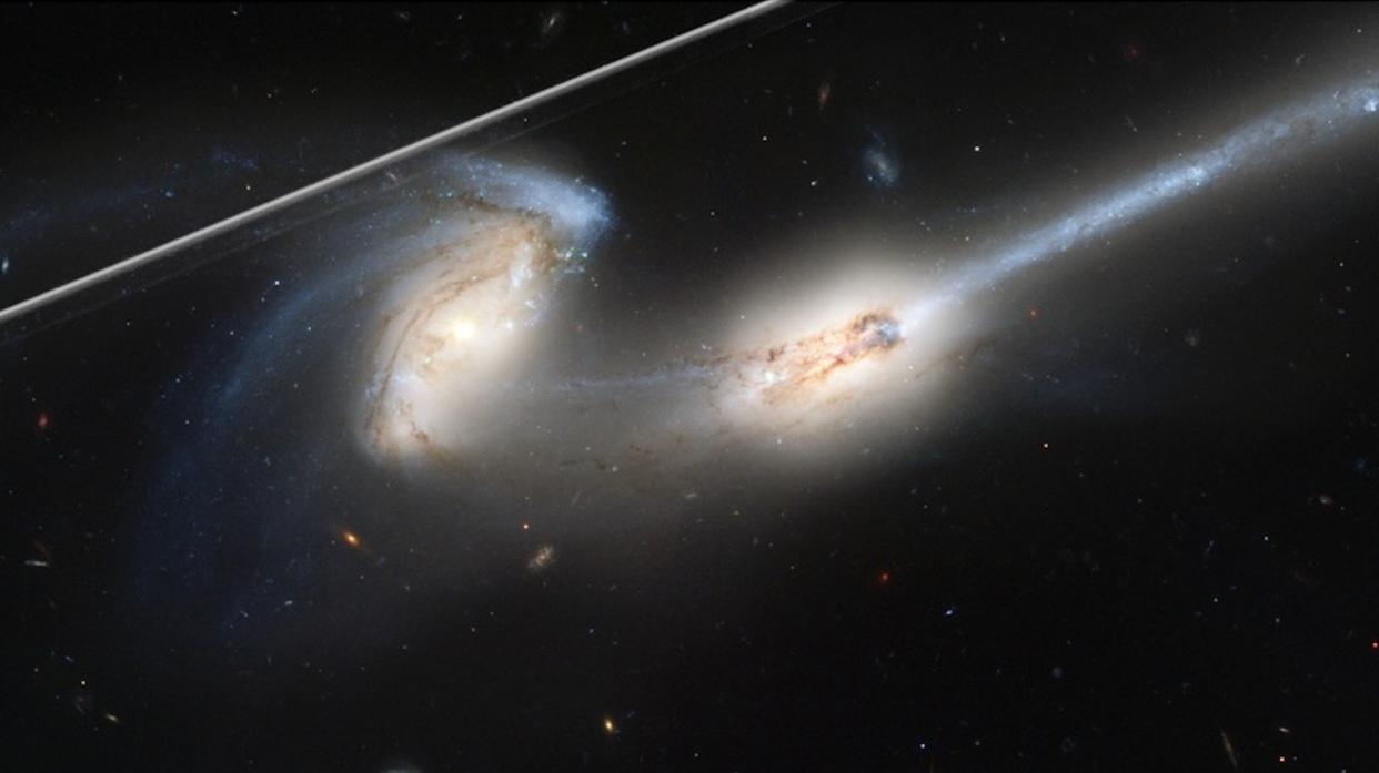  Hubble photo of two galaxies in deep space marred by a satellite trail, which appears as a bright white line across the upper left of the image. 