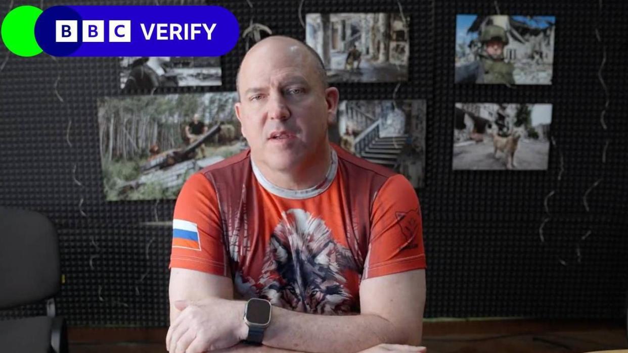 A bald man looking at the camera wearing a t-shirt with a wolf on the front and a Russian flag on the sleeve