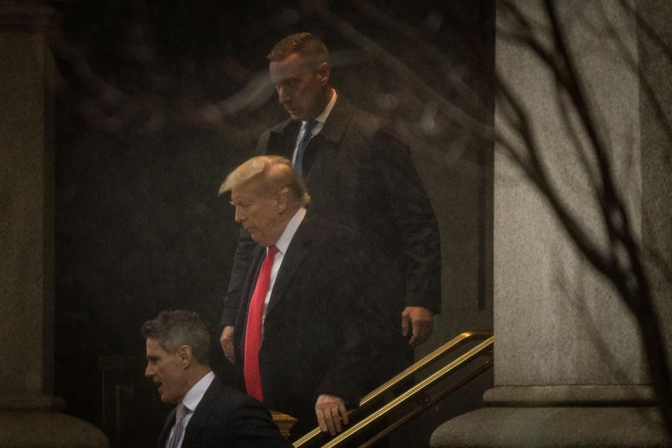 Donald Trump wears a black jacket and red tie and walks down steps outside, flanked by two men also in suits.