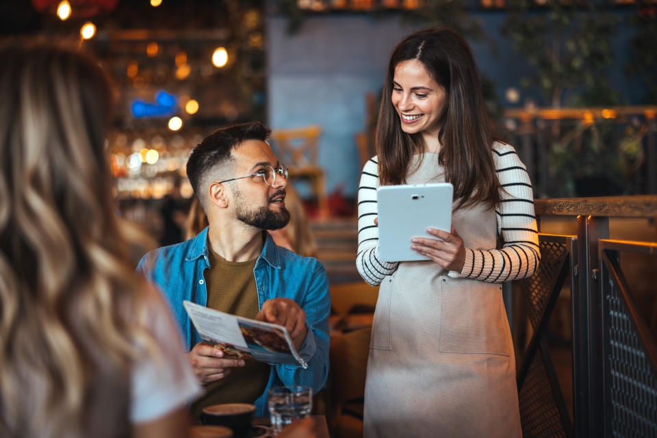 Two unidentified people smile and discuss a menu in a casual dining setting; one holds a newspaper, the other a tablet