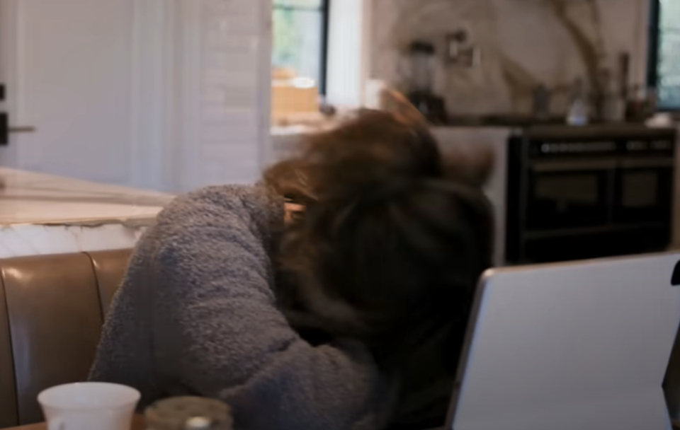 Jennifer Lopez resting head on table next to laptop, appears frustrated or tired