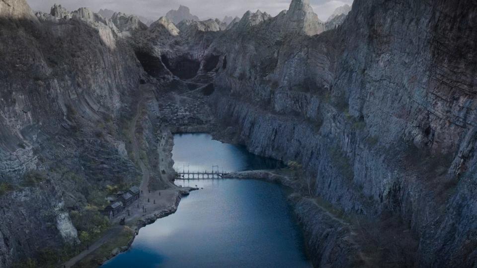 A wide, establishing shot of a mountain valley dominated by a lake and three huge pits at the far end, clearly a mine. There is a ramshackle village in front of the mine, and the lake is spanned by a rickety bridge.