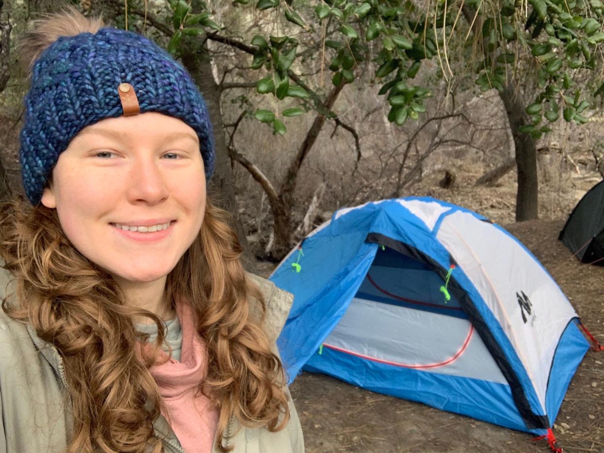 woman with curly hair wearing blue hat selfie smiling in front of tent