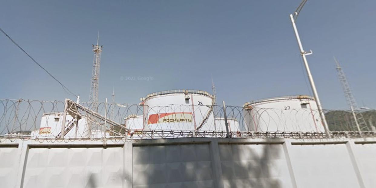 A Google streetview image of a Rosneft Tuapse oil depot in Tuapse, Russia, dated to 2012. A tall concrete wall topped by barbed wire partially blocks the view of several white containers, against a blue sky.