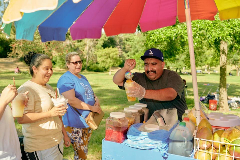 Three people wait as a man serves up raspado under a colored umbrella.