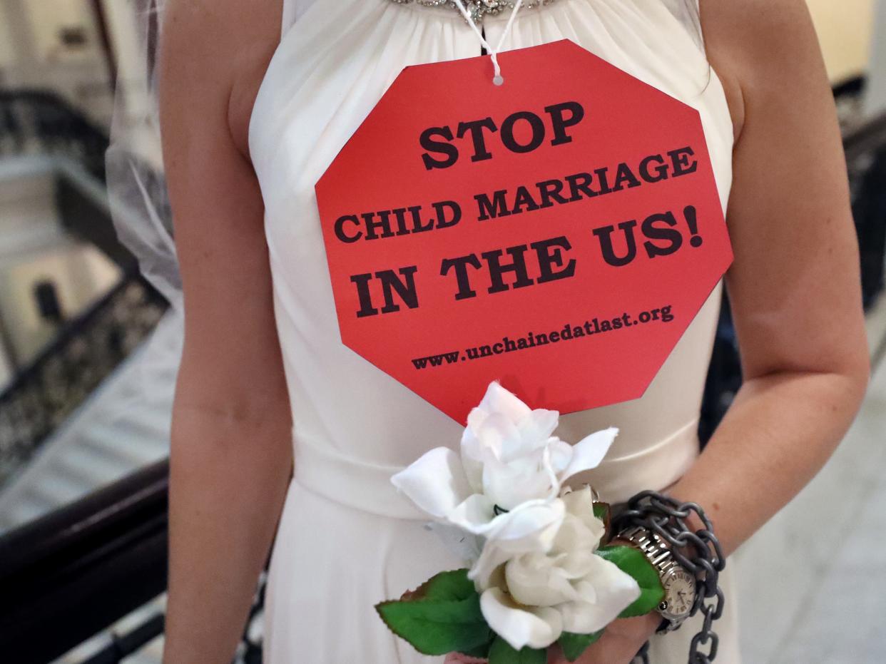 A woman in a white dress is shown from the neck down with a red stop sign-shaped sign reading "Stop child marriage in the US!" She is holding flowers and has a chain wrapped around her wrist.