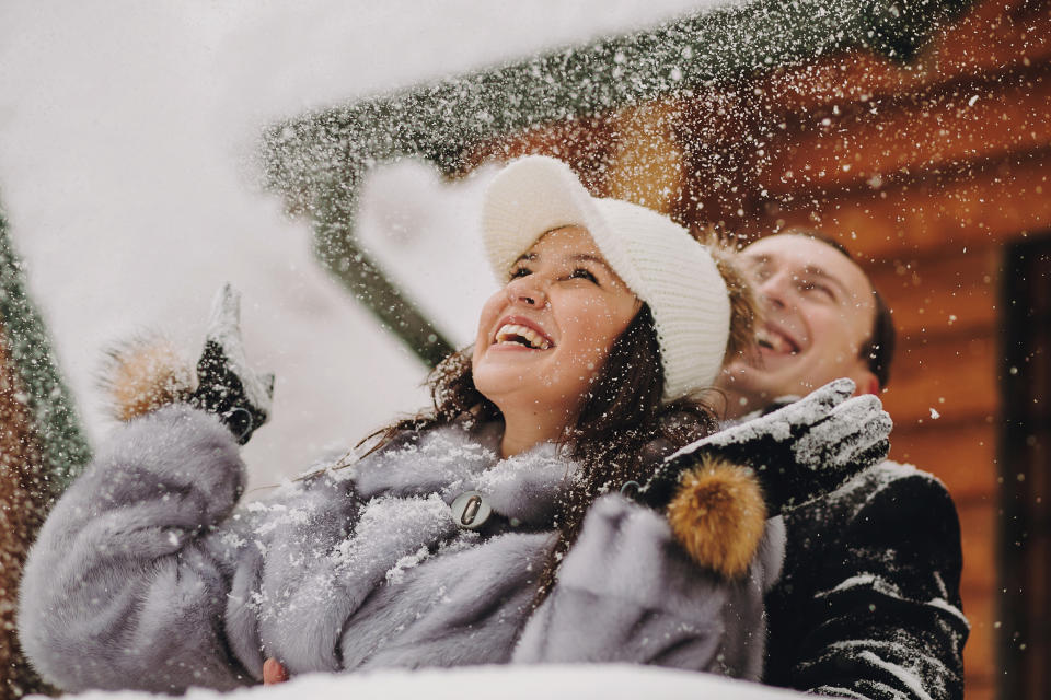 Stylish couple playing with snow in wooden cabin on background of winter snowy mountains. Happy joyful family having fun and smiling in snow. Emotional funny moments together