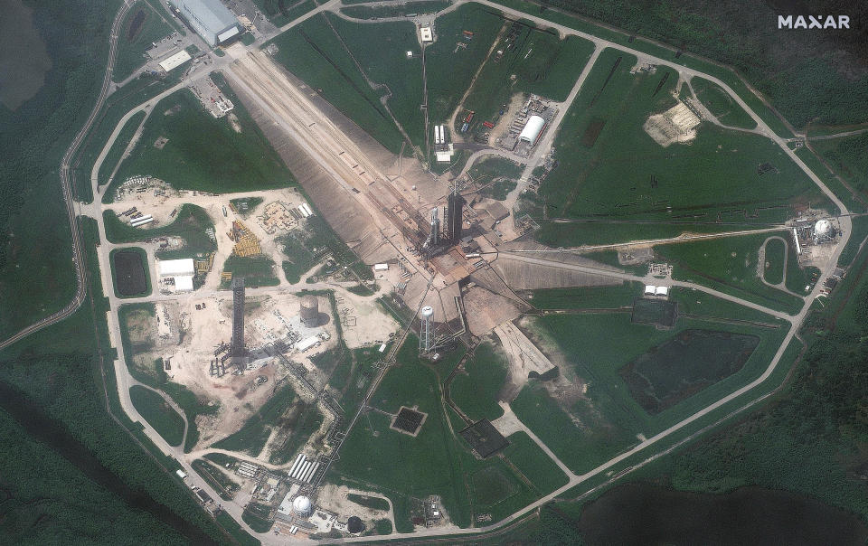 A high-altitude aerial view of SpaceX's triple-booster Falcon Heavy rocket can be seen standing in the center of the wide landscaped launch complex. Roads, pipes and other infrastructure extend outward from the rocket and adjacent launch tower.