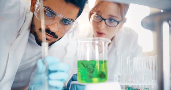 Two scientists in lab in front of a beaker containing green fluid.