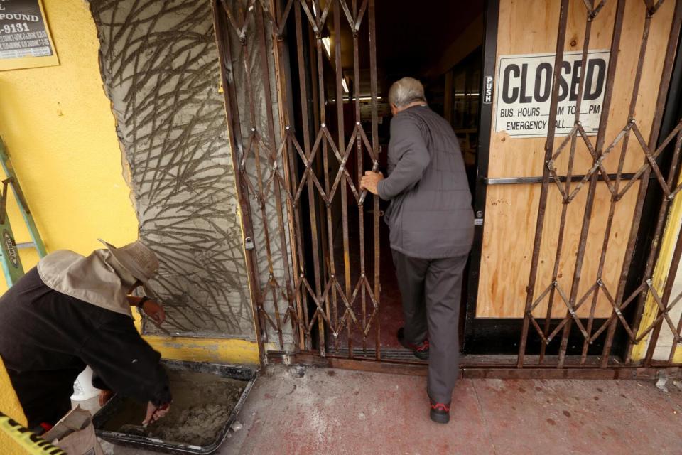 A man, seen from behind, opens a folding exterior metal door as he enters a business with a boarded-up front door
