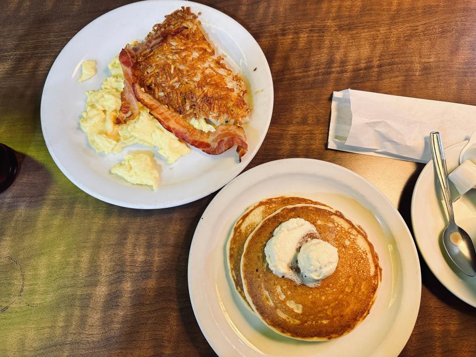 Two plates of Denny's breakfast, the one on the left with eggs, hash browns, and bacon, and the one on the right with two pancakes