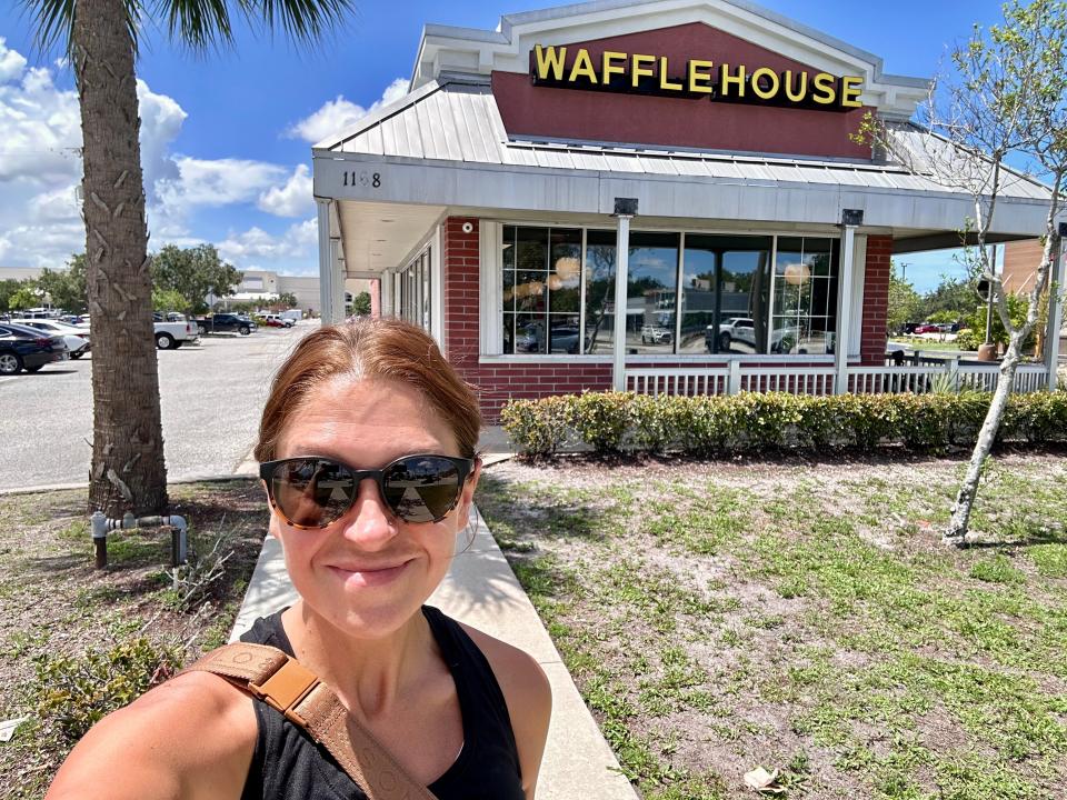 Selfie of the writer in front of Waffle House. The building is red with a white roof and yellow lettering on it.