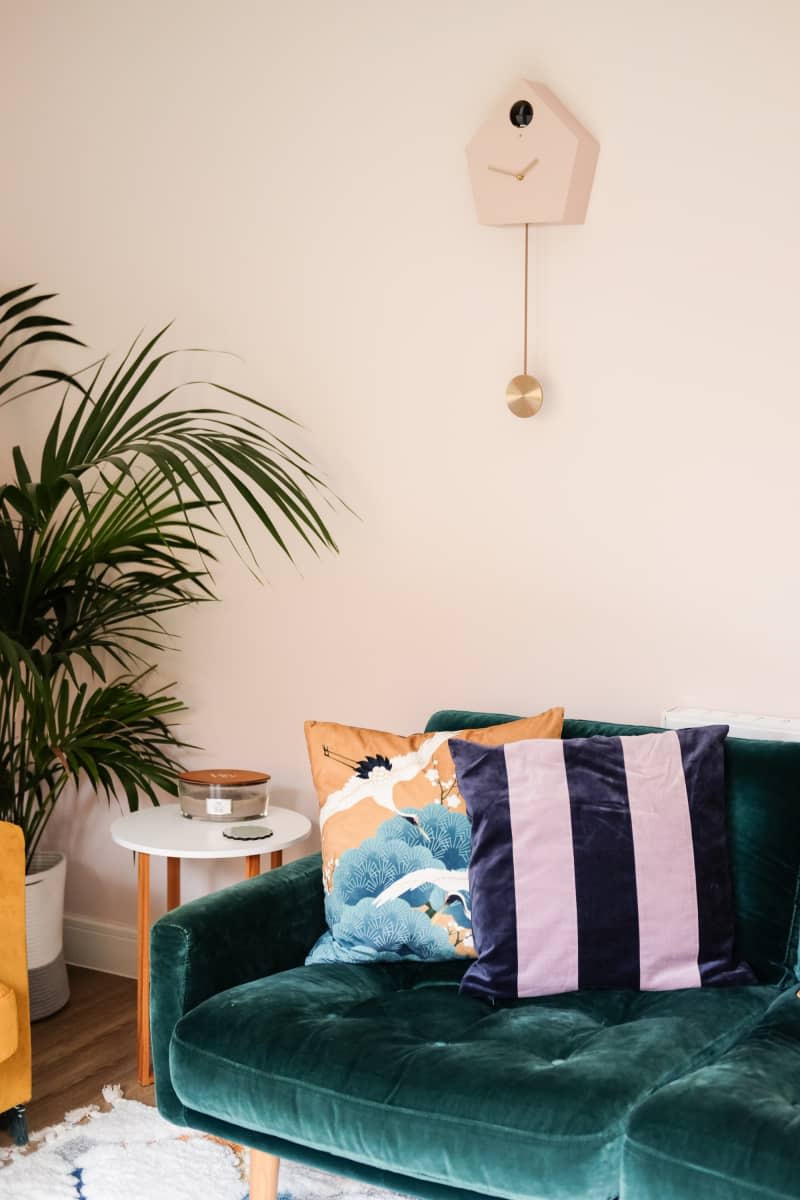 teal velvet couch with pink and black striped throw pillow and light pink cuckoo clock on wall, large fern in corner