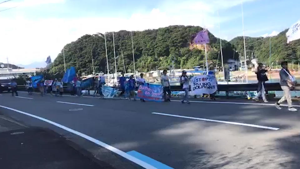 A protest march on the other side of a narrow Japanese road. Hills and water in the background. 