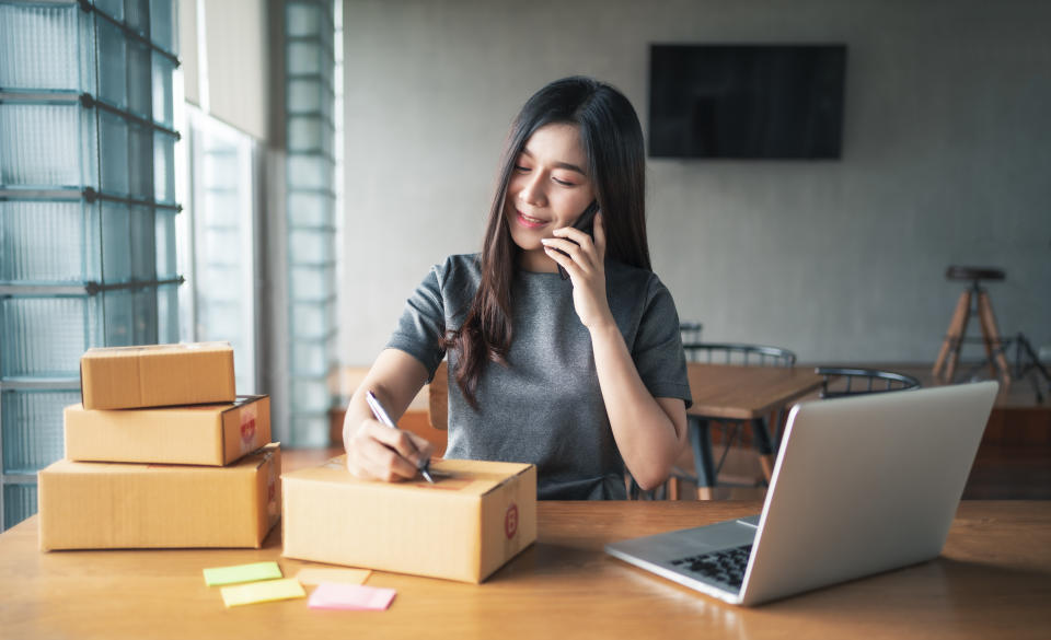 Young business woman working selling online. Entrepreneur owner using smartphone or laptop taking receive and checking online purchase shopping order to preparing pack product box. Packing goods for delivery to customer. Online selling. E-commerce. Online Shopping