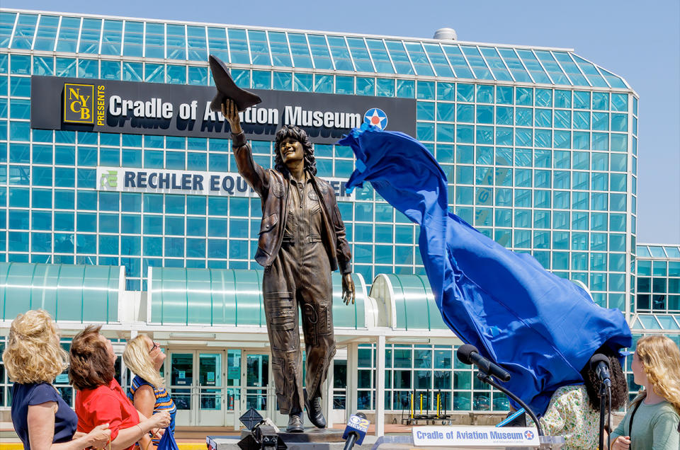 a dark-brown statue of a woman holding a small space shuttle stands in front of a bluish-green building