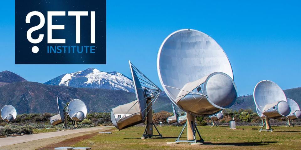 a large white satellite dish sits in a grass field in front of a large snowy mountain against a blue sky background.