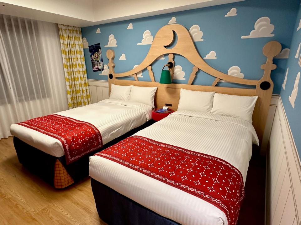 The guest room at Tokyo's Toy Story Hotel features an oversized headboard with two twin beds with white sheets and a red blanket. Blue wallpaper with white clouds covers the walls.