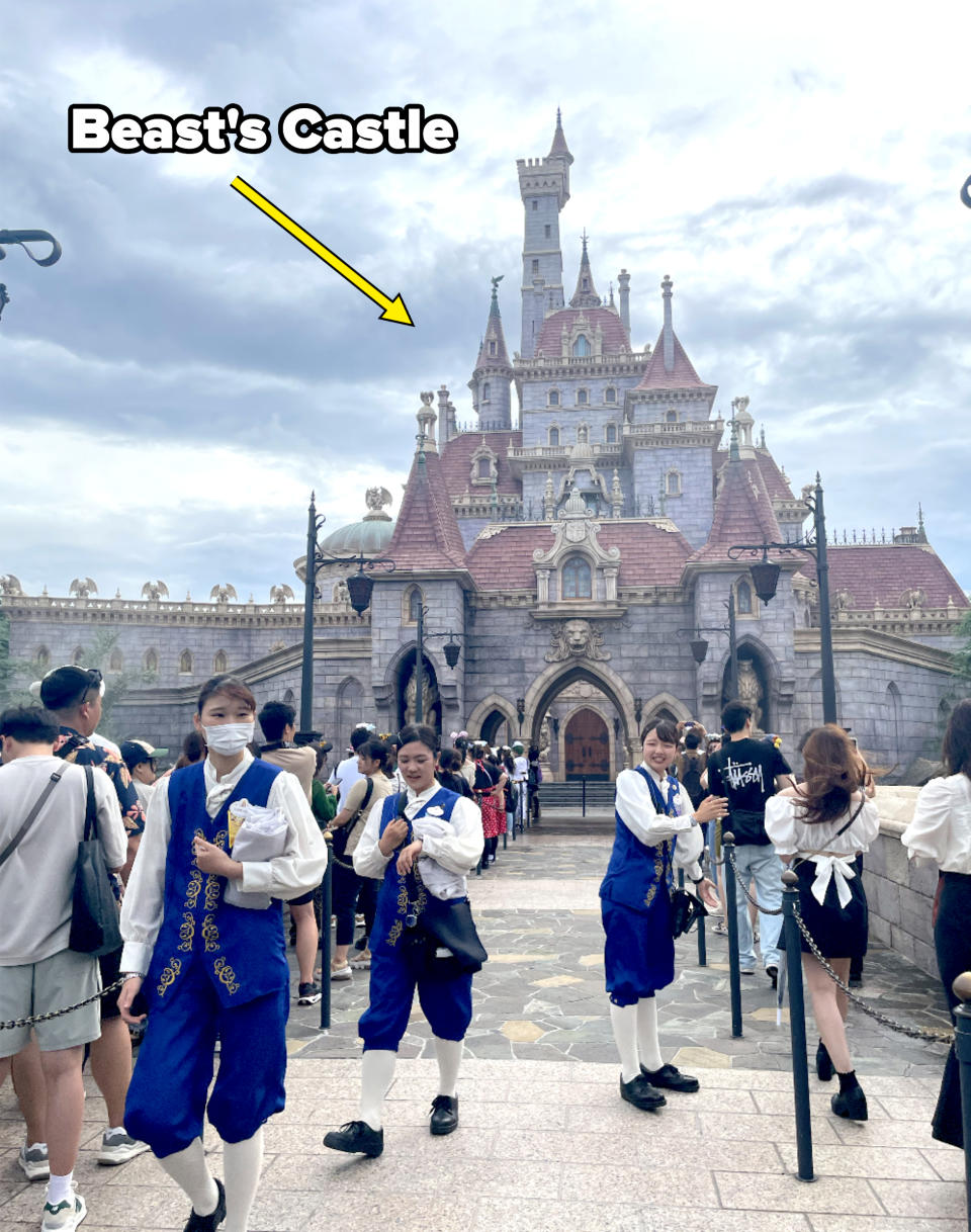 People dressed as Disney cast members in themed uniforms stand in front of the Beast's Castle at Tokyo Disneyland, surrounded by visitors