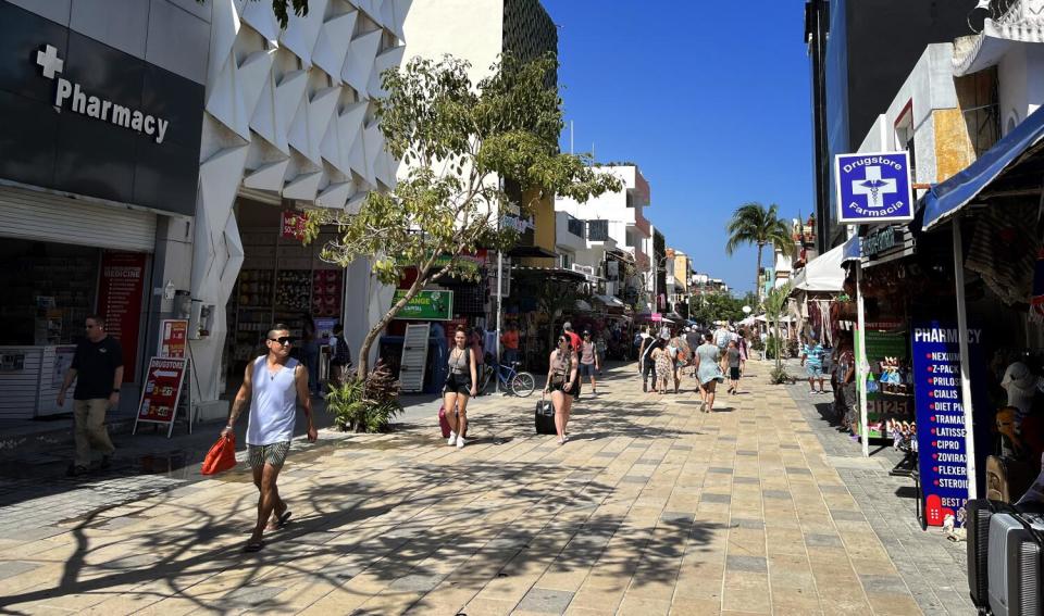 People walk along a pedestrian street lined by buildings, including two pharmacies right across from each other