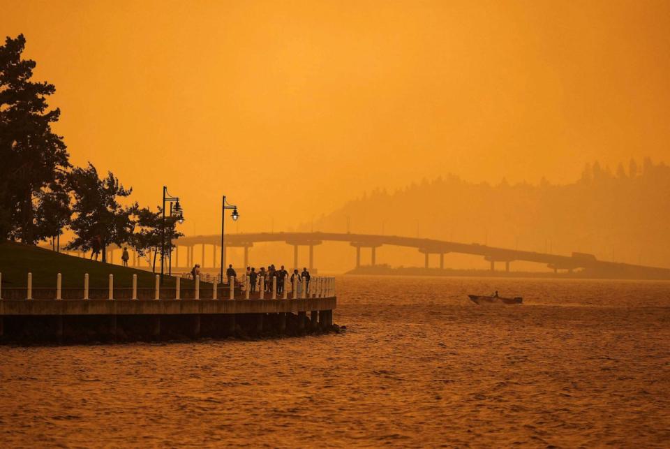 PHOTO: A person travels in a boat past people walking on the boardwalk as smoke from the McDougall Creek wildfire blankets the area on Okanagan Lake in Kelowna, British Columbia, Aug. 18, 2023. (Darryl Dyck/AP)