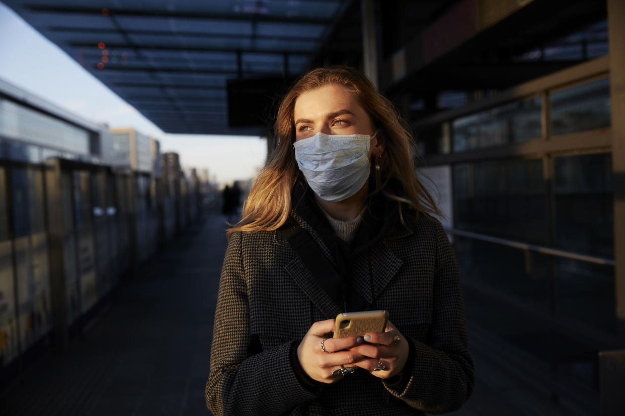 Young woman standing on train station wearing protective mask, using phone