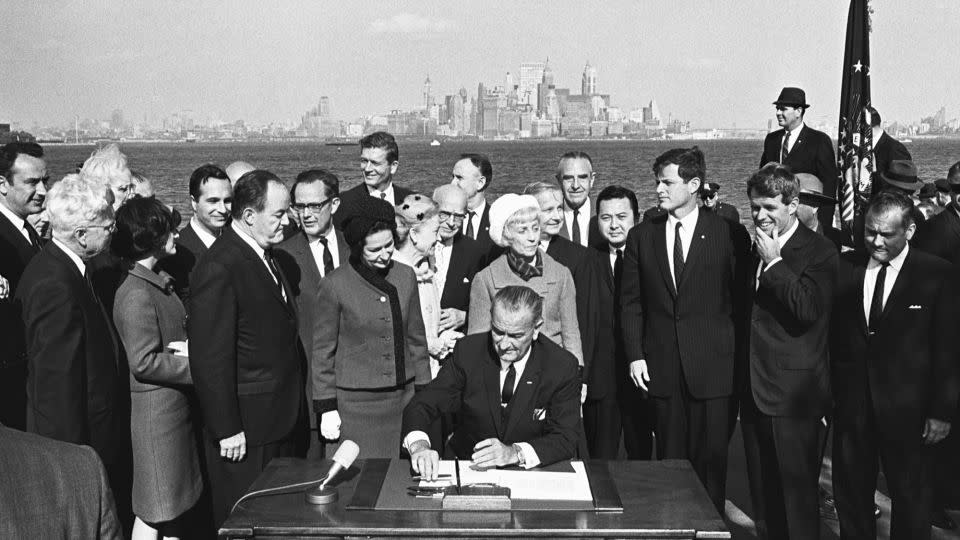 President Lyndon B. Johnson signs the Immigration Bill of 1965 on Liberty Island in New York Harbor with a view of the New York City skyline in the background. Next to the president on his right are First Lady Lady Bird Johnson and Vice President Hubert Humphrey. To the president's left are Senator Edward Kennedy (third from right) and Robert Kennedy (second from right). - Corbis Historical/Getty Images