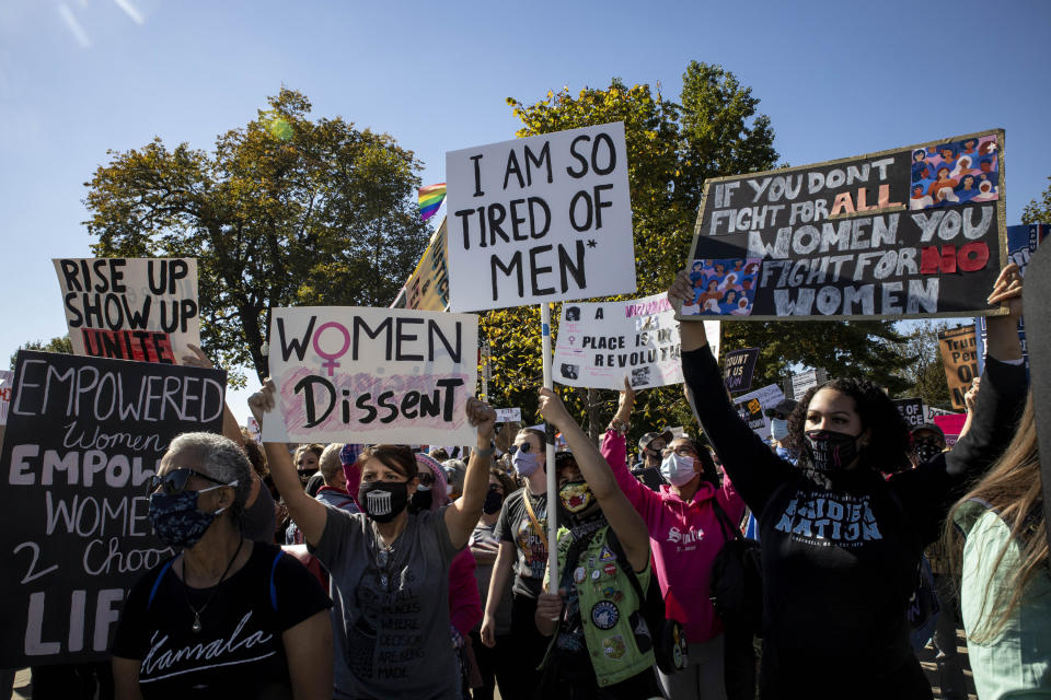 Protesters stand in a park and hold up signs reading "I am so tired of men" and "women dissent" and "if you don't fight for all women you fight for no women"