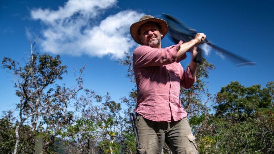 man in wide-brimmed hat spinning a butterfly net with forest and blue sky in the background