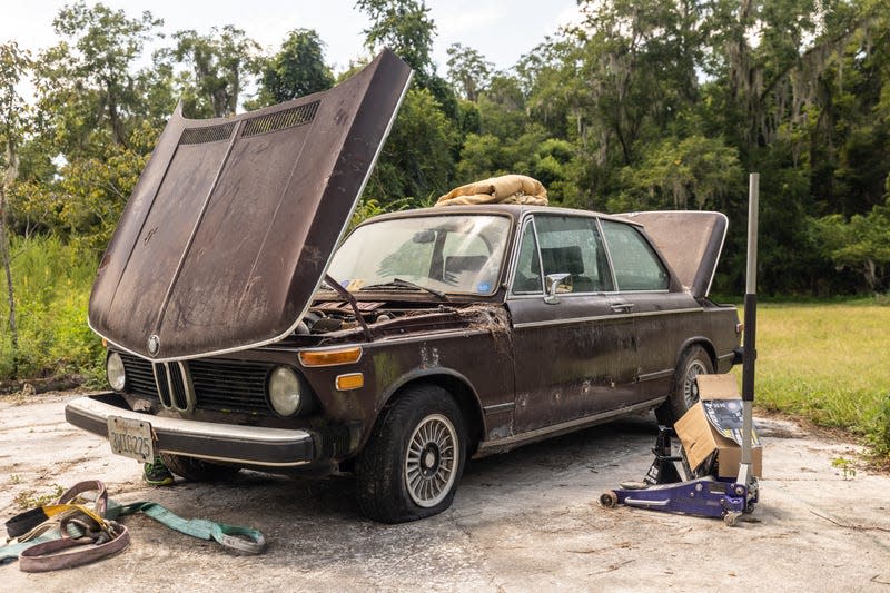 a maroon 1974 bmw 2002tii sits on a concrete pad in a field with its hood and trunk open. the car has been abandoned for decades; it's covered in dirt and grime and surrounded by tools, jacks, and tow straps, all of which were used to pull it out of the field where it sat for 25 years