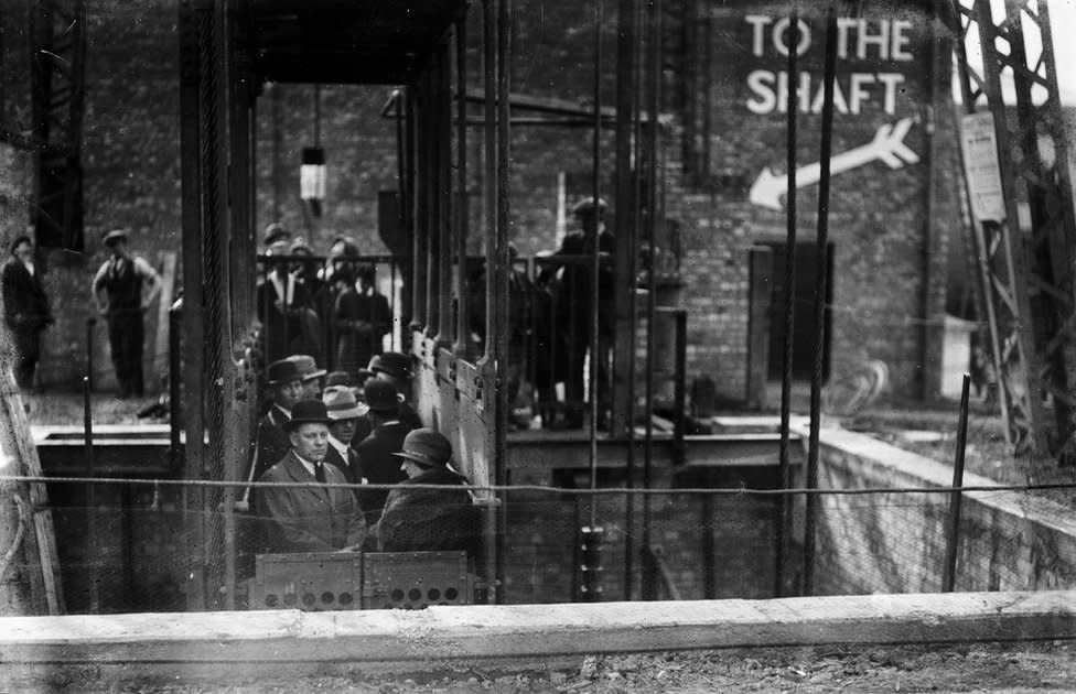 Black and white image of people standing in cage descending into the mine shift display