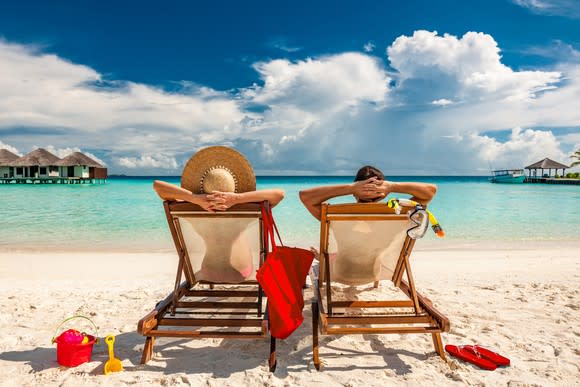 Two people in recliners on a beach with their hands behind their heads. The sky is blue, the water is clear, and there are some clouds and sand.